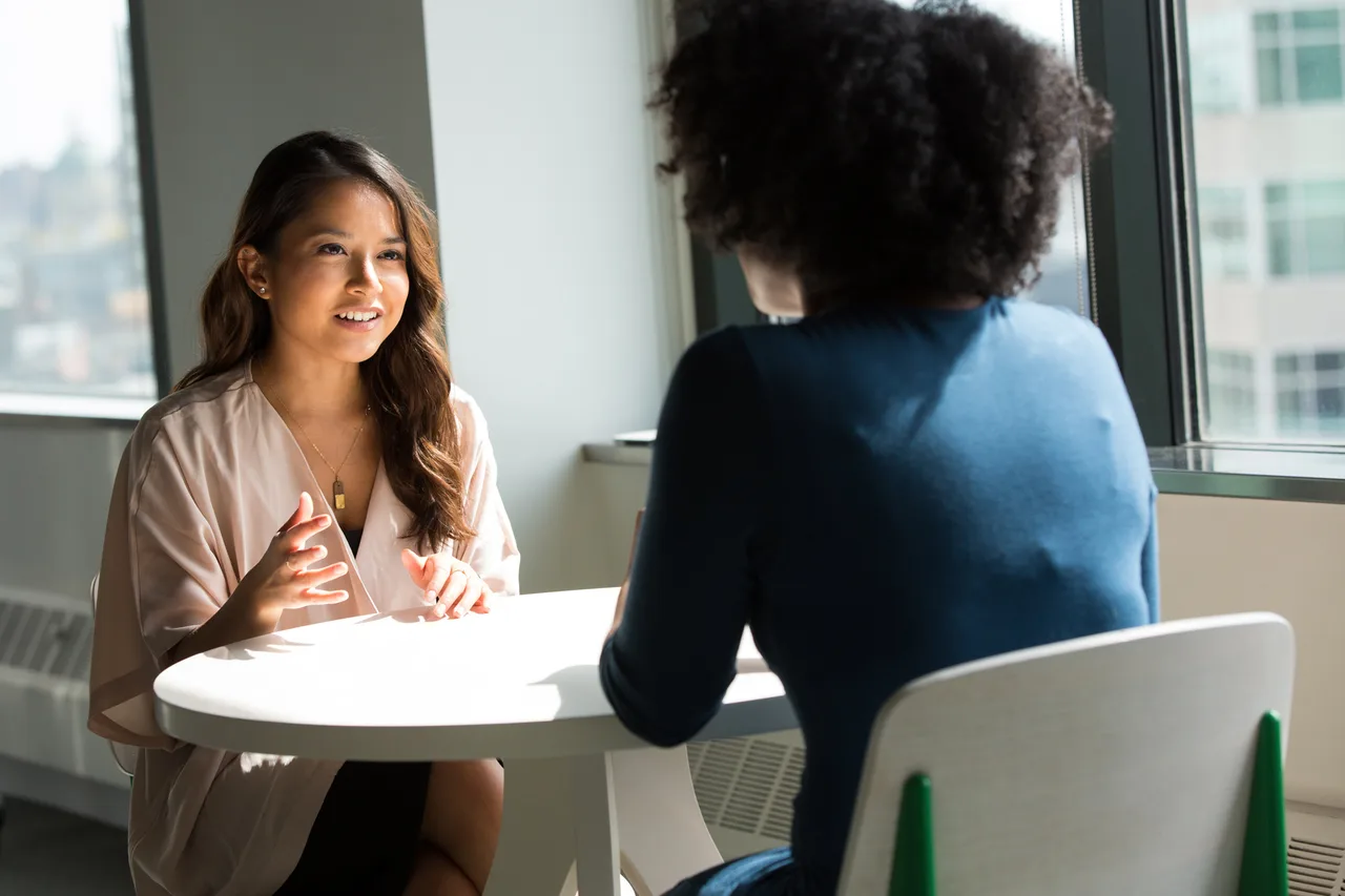 Two women talking at a round white table in a bright office with a city view.