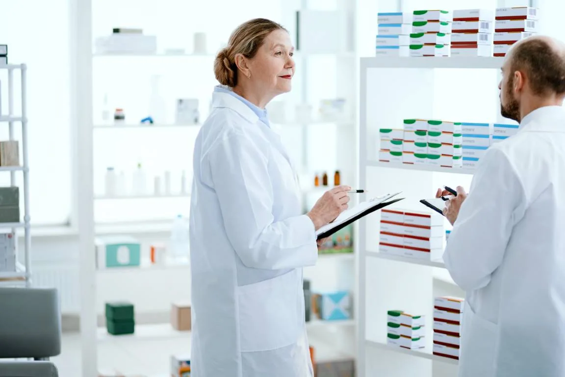 Two scientists in white coats talking in a laboratory, shelves of coloured flasks and bar charts on the wall.